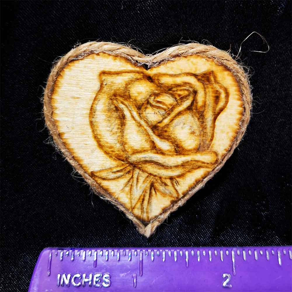 alt="Horizontal ruler measuring a small 2 inch by 2 inch hand burned heart shaped wood slice magnet with a single rose art finished with a brown twine edge border.">