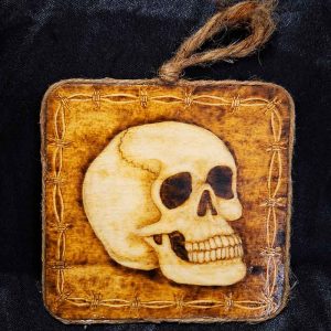 alt="4 inch by 4 inch square wood slice with hand burned skull art with a barbed wire border finished with a brown twine outer edge with a loop for wall hanging.">
