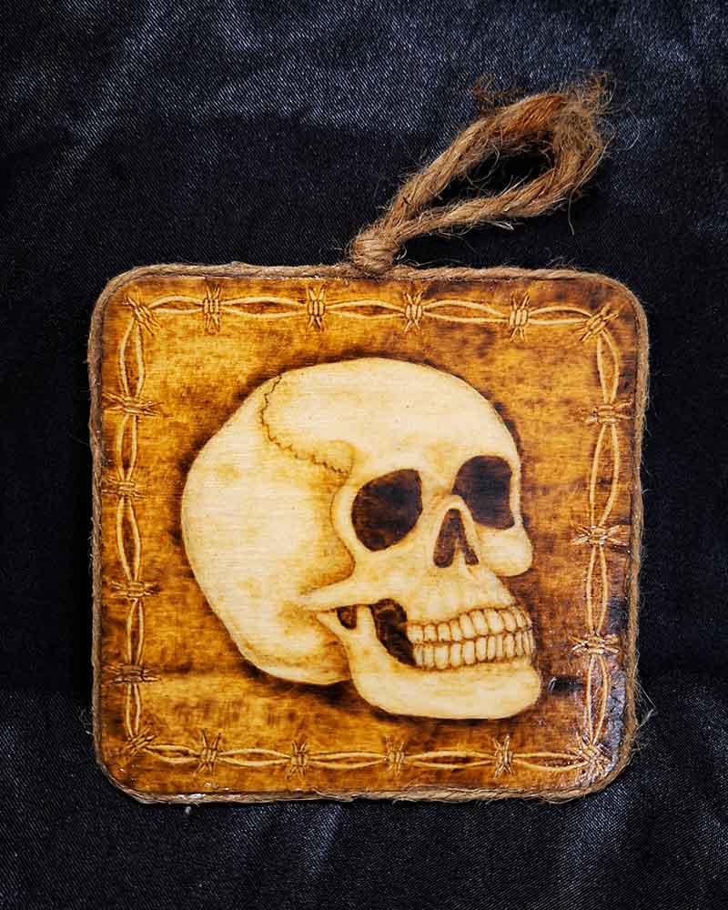 alt="4 inch by 4 inch square wood slice with hand burned skull art with a barbed wire border finished with a brown twine outer edge with a loop for wall hanging.">