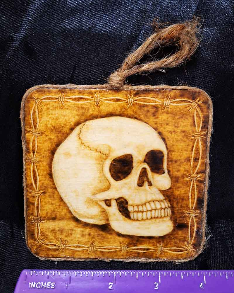 alt="Horizontal ruler measuring a 4 inch by 4 inch square wood slice with hand burned skull art with a barbed wire border finished with a brown twine outer edge with a loop for wall hanging.">