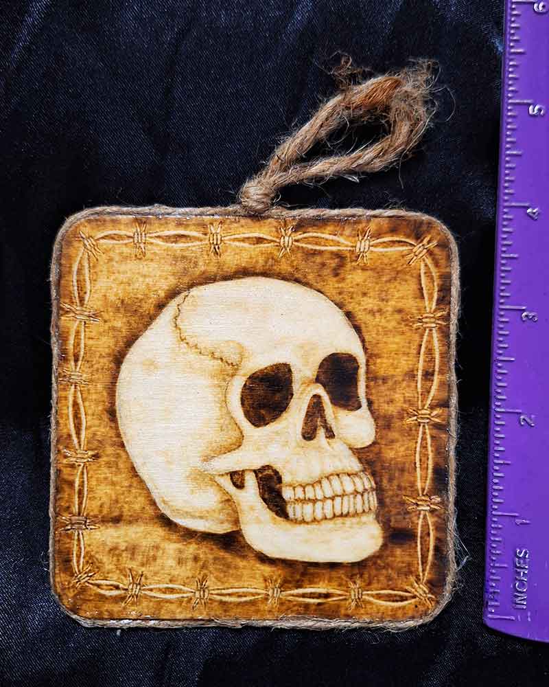 alt="Vertical ruler measuring a 4 inch by 4 inch square wood slice with hand burned skull art with a barbed wire border finished with a brown twine outer edge with a loop for wall hanging.">