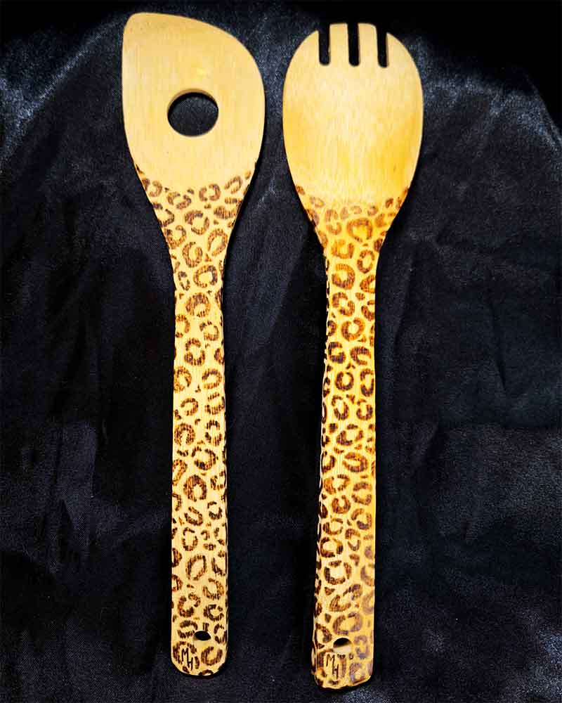 alt="Two side by side full size wooden kitchen utensils, one slotted end spoon and one pasta hole spatula designed with hand burned leopard print fully wrapping the entire handle.">