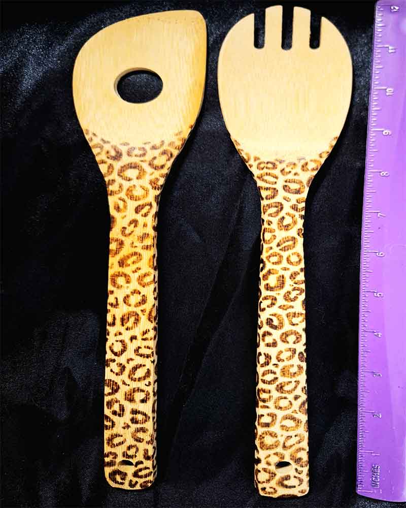alt="Vertical ruler measuring two side by side full size wooden kitchen utensils, one slotted end spoon and one pasta hole spatula designed with hand burned leopard print fully wrapping the entire handle.">