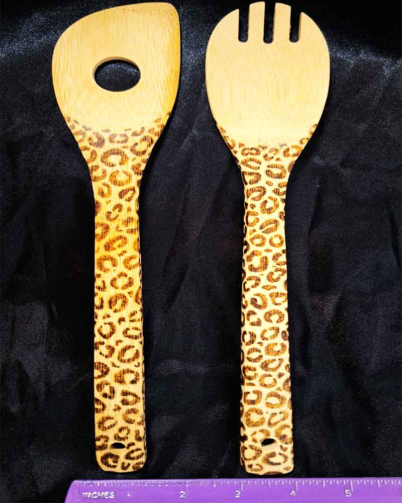 alt="Horizontal ruler measuring two side by side full size wooden kitchen utensils, one slotted end spoon and one pasta hole spatula designed with hand burned leopard print fully wrapping the entire handle.">