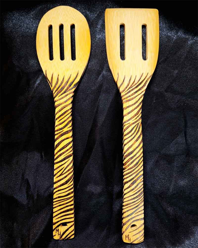 alt="Two side by side full size wooden kitchen utensils, one slotted spoon and one slotted spatula designed with hand burned zebra stripes fully wrapping the entire handle.">