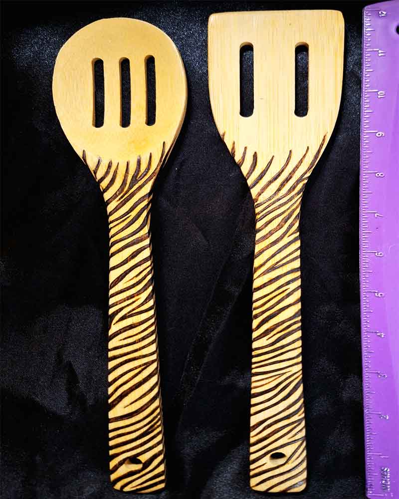alt="Vertical ruler measuring two side by side full size wooden kitchen utensils, one slotted spoon and one slotted spatula designed with hand burned zebra stripes fully wrapping the entire handle.">
