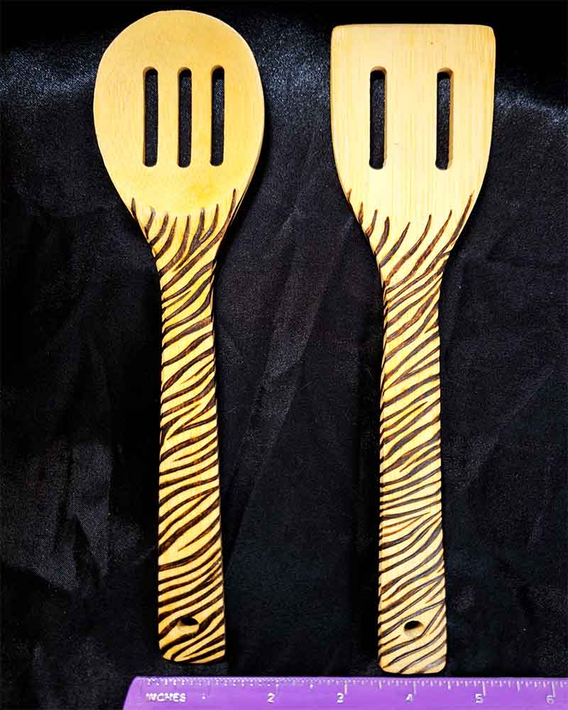 alt="Horizontal ruler measuring two side by side full size wooden kitchen utensils, one slotted spoon and one slotted spatula designed with hand burned zebra stripes fully wrapping the entire handle.">