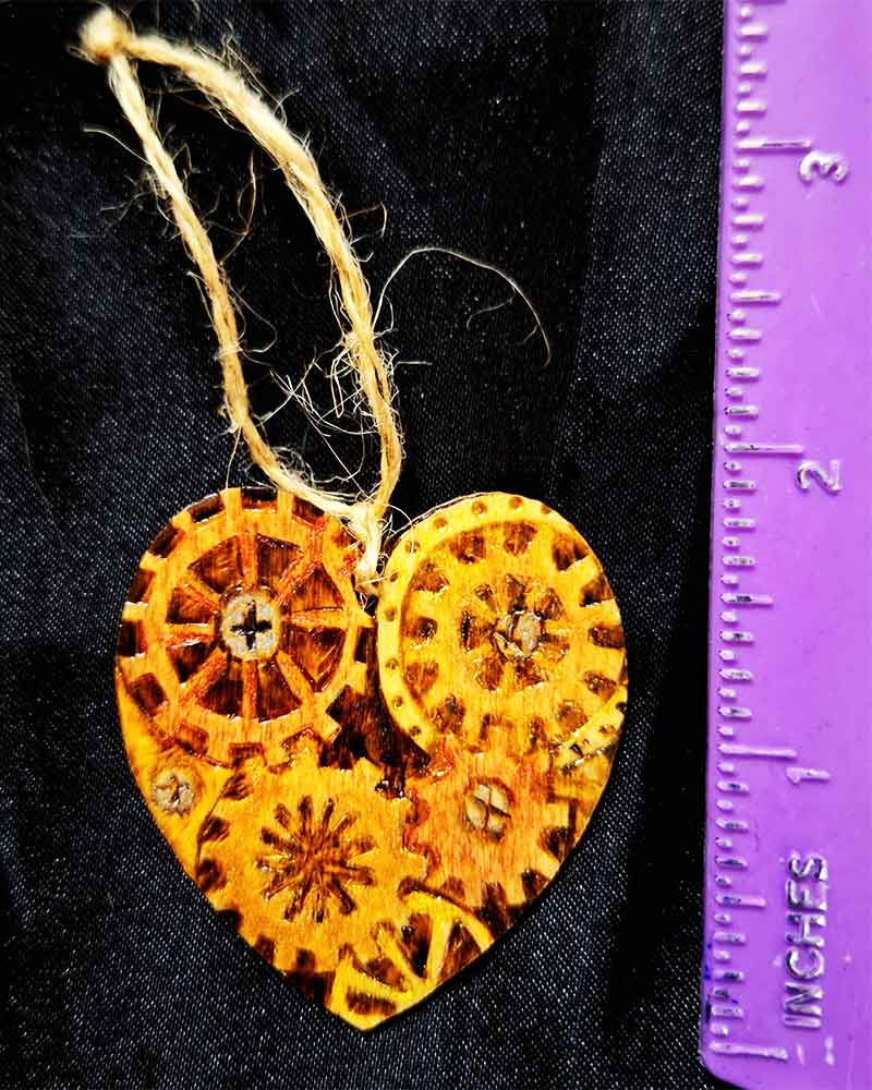 alt="Vertical ruler measuring the front of a small 2 inch by 2 inch hand burned heart shaped wood slice with steampunk art with metallic painted gears finished with a brown twine loop for wall hanging.">