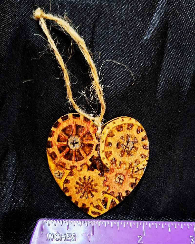 alt="Horizontal ruler measuring the front of a small 2 inch by 2 inch hand burned heart shaped wood slice with steampunk art with metallic painted gears finished with a brown twine loop for wall hanging.">