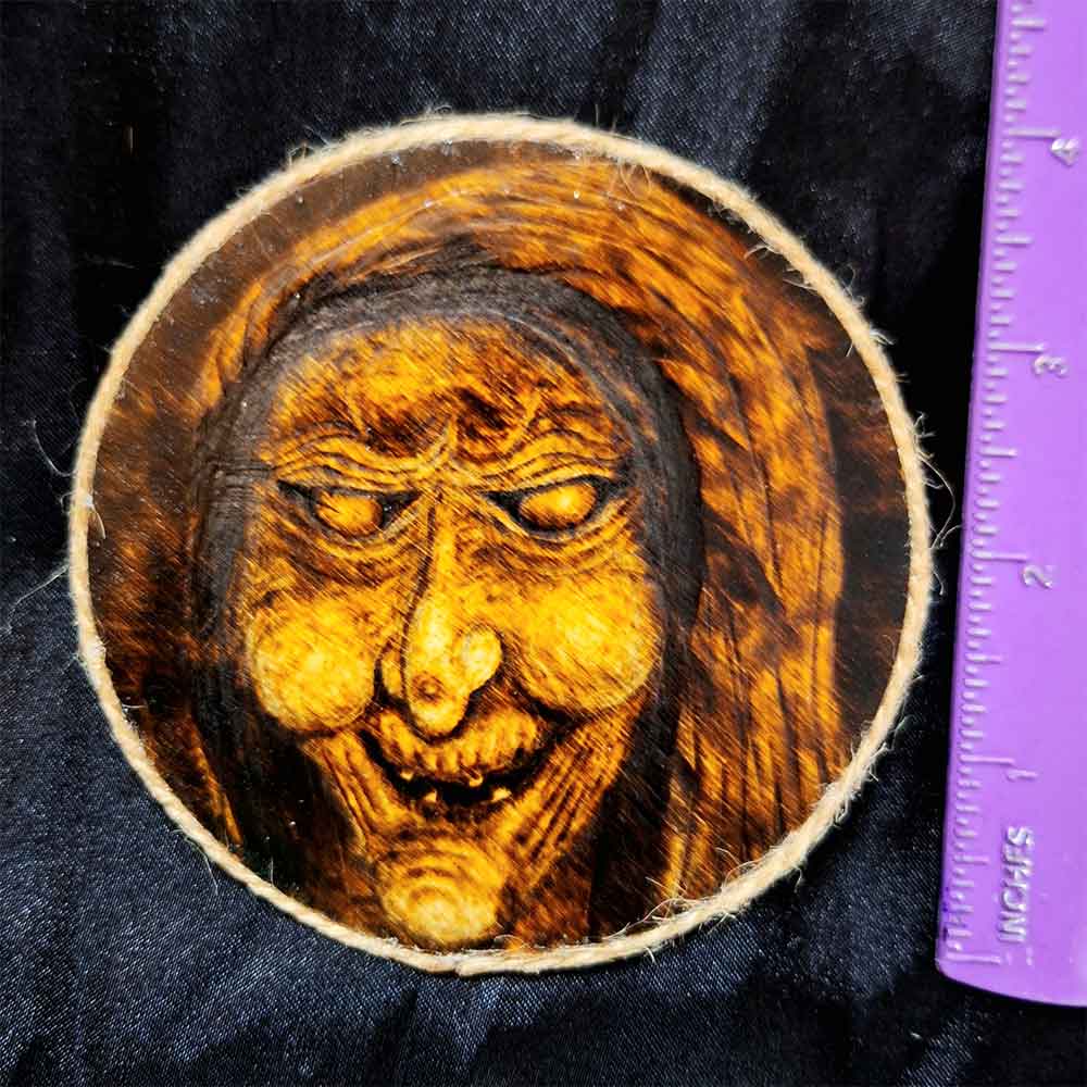alt="Vertical ruler measuring a 4 inch by 4 inch round wood slice magnet designed with hand burned haunting witch hag artwork finished with a brown twine outer edge.">
