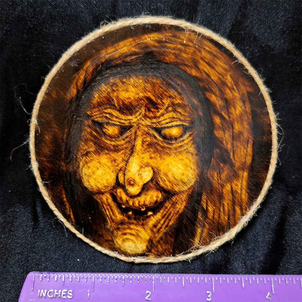 alt="Horizontal ruler measuring a 4 inch by 4 inch round wood slice magnet designed with hand burned haunting witch hag artwork finished with a brown twine outer edge.">