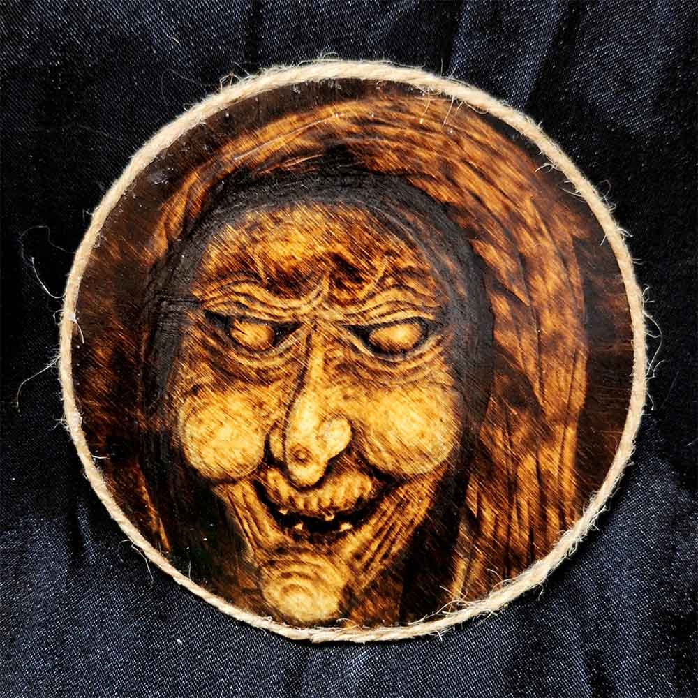 alt="4 inch by 4 inch round wood slice magnet designed with hand burned haunting witch hag artwork finished with a brown twine outer edge.">